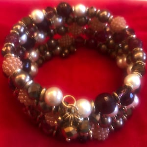May include: A multi-strand wrap bracelet featuring a mix of beads in various colors and textures. The bracelet includes white faux pearls, dark red, brown, and gold-colored beads, and small, textured, peach-colored beads. The bracelet is set against a red background.