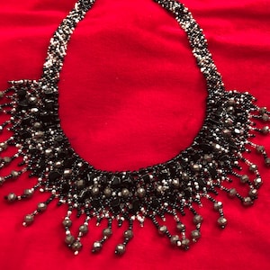 May include: A beaded necklace with a bib design. The necklace features black, silver, and white beads. The bib portion has dangling bead accents. The necklace is displayed on a red surface.
