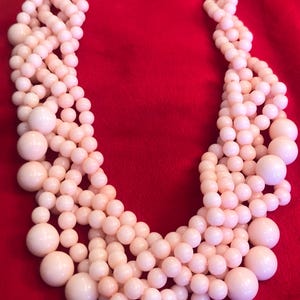 May include: A multi-strand necklace featuring light pink beads of varying sizes. The necklace has a gold-colored clasp and is displayed against a red background. The beads range from small to large, creating a layered effect.