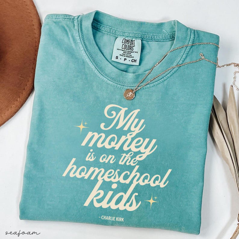 May include: Seafoam green t-shirt with the text "My money is on the homeschool kids" in a stylized font. A gold necklace with a pendant rests on the shirt. The shirt has a Comfort Colors tag.