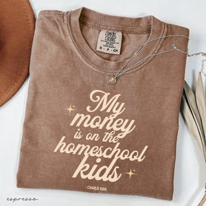 May include: Espresso-colored Comfort Colors t-shirt with the text "My money is on the homeschool kids" in a retro font. A silver necklace with a pendant rests on the shirt. The shirt is size S-P-CH.