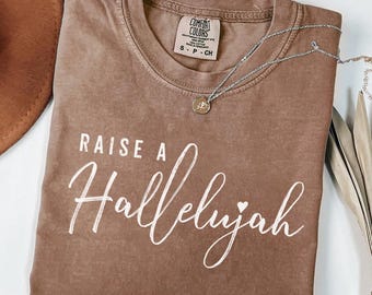 May include: A brown Comfort Colors t-shirt with the phrase "RAISE A Hallelujah" in white script. A silver necklace with a small pendant rests on the shirt. The shirt is folded and displayed with a brown hat.