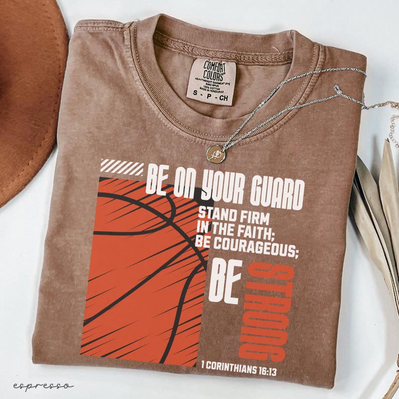 May include: Espresso-colored t-shirt with the text "BE ON YOUR GUARD" and a basketball graphic. The shirt also features the text "STAND FIRM IN THE FAITH; BE COURAGEOUS; BE STRONG" and "1 CORINTHIANS 16:13". A silver necklace is draped across the shirt.