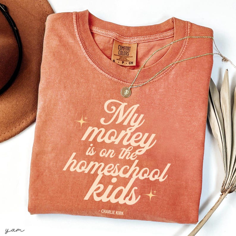 May include: A coral-colored Comfort Colors t-shirt with the text "My money is on the homeschool kids" in a stylized font. A gold necklace with a pendant is draped across the shirt. A brown hat is partially visible.