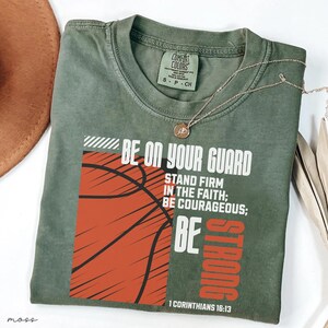 May include: Olive green t-shirt with a basketball graphic and the text "BE ON YOUR GUARD". The shirt also features the text "STAND FIRM IN THE FAITH; BE COURAGEOUS; BE STRONG" and "1 CORINTHIANS 16:13". A gold necklace is draped across the shirt.