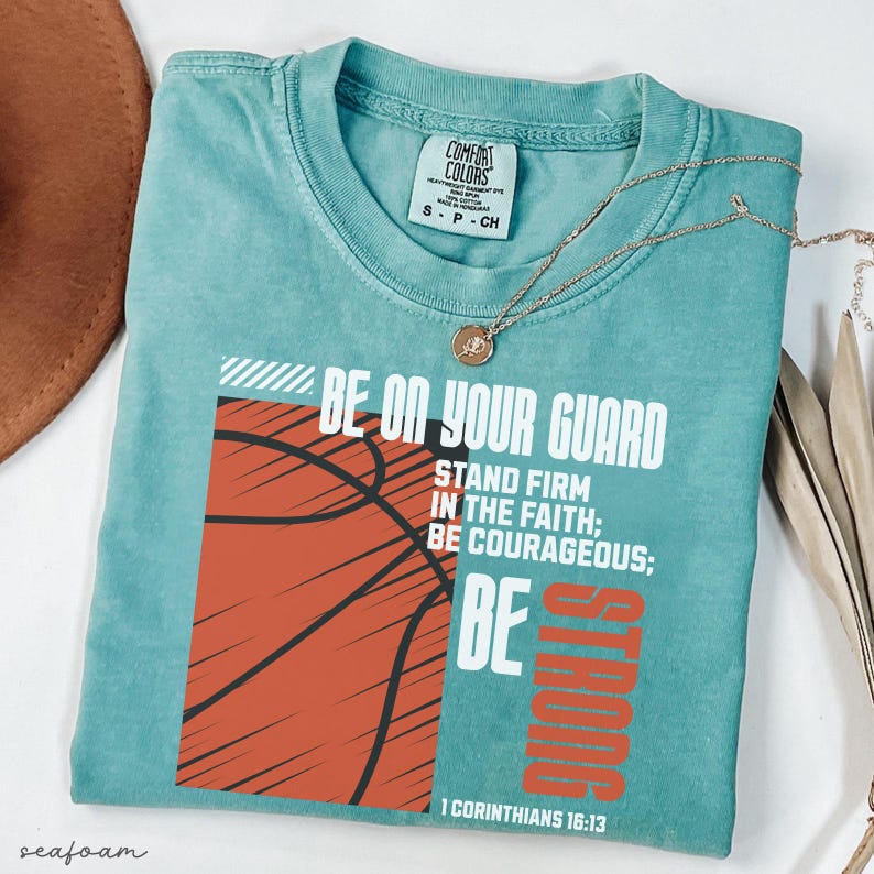 May include: Seafoam green t-shirt with a basketball graphic and the text "BE ON YOUR GUARD, STAND FIRM IN THE FAITH; BE COURAGEOUS; BE STRONG" in white and orange. A gold necklace with a pendant is on the shirt.