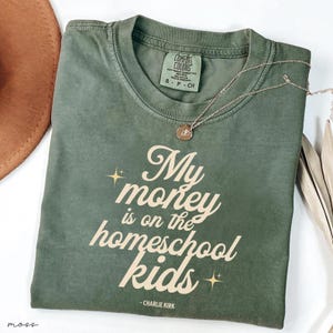 May include: An olive green t-shirt with the text "My money is on the homeschool kids" in a cursive font. The shirt has a crew neck and short sleeves. A gold necklace with a pendant is draped across the shirt.