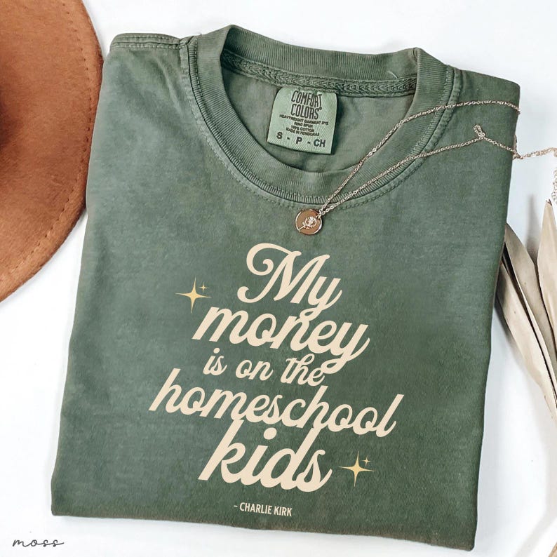 May include: An olive green t-shirt with the text "My money is on the homeschool kids" in a cursive font. The shirt has a crew neck and short sleeves. A gold necklace with a pendant is draped across the shirt.