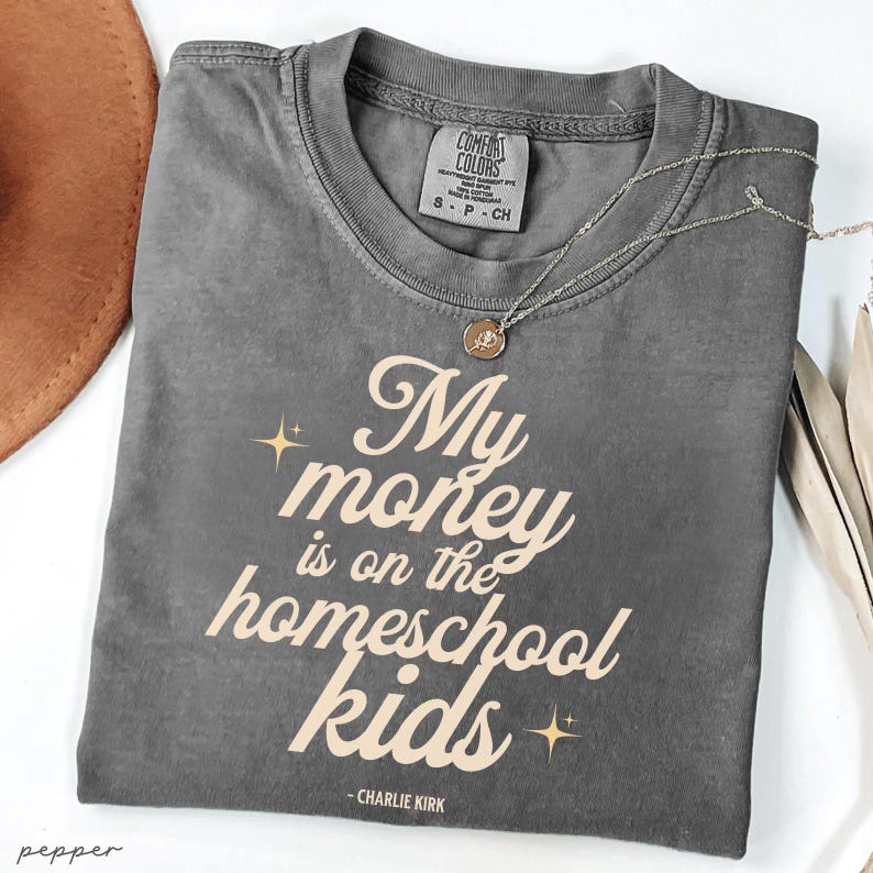May include: A gray Comfort Colors t-shirt with the text "My money is on the homeschool kids" in a light beige color. A silver necklace with a pendant is on the shirt. A brown hat is visible in the background.