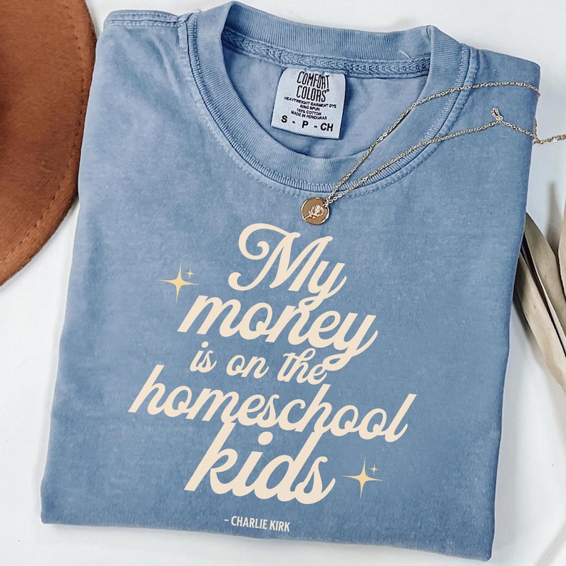 May include: A blue Comfort Colors t-shirt with the text "My money is on the homeschool kids" in a cream-colored font. A gold necklace with a pendant is draped across the shirt. The shirt also has the text "- CHARLIE KIRK".