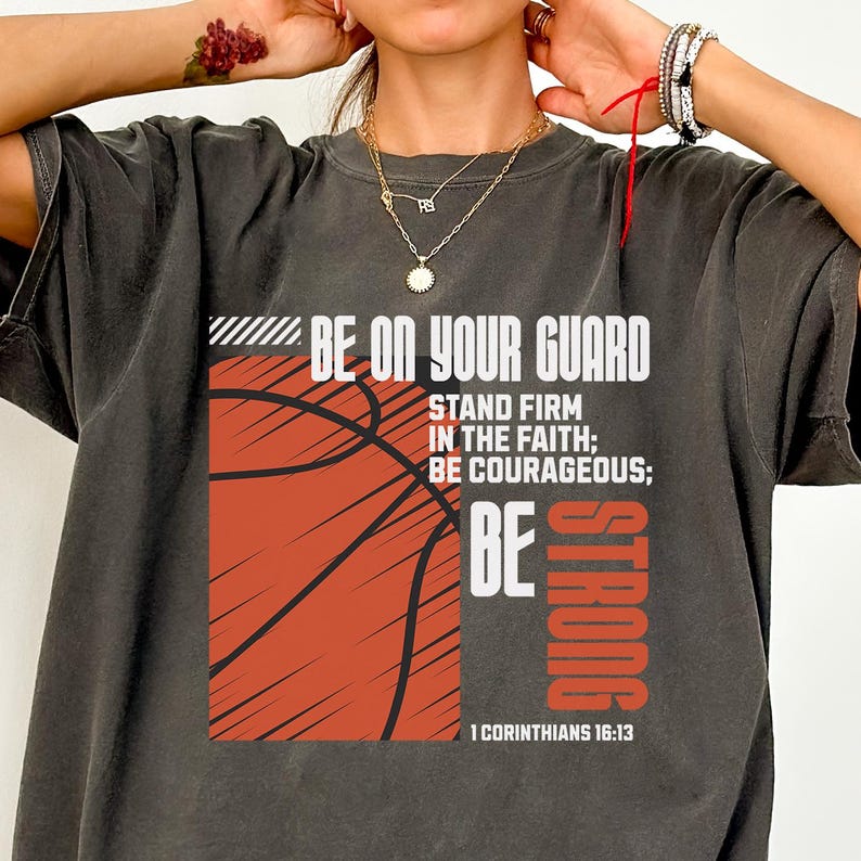 May include: Dark gray t-shirt with a basketball graphic and the text "BE ON YOUR GUARD". The design includes the words "STAND FIRM IN THE FAITH; BE COURAGEOUS; BE STRONG" and "1 CORINTHIANS 16:13".