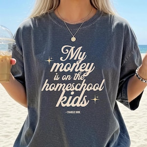 May include: Dark gray t-shirt with the text "My money is on the homeschool kids" in a white, stylized font. The quote is attributed to Charlie Kirk. The shirt has a relaxed fit and short sleeves.