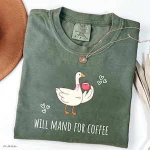 Puede incluir: Camiseta verde oliva con un dibujo de un ganso sosteniendo una taza de café, con el texto "WILL MAND FOR COFFEE". Un collar plateado con un colgante está sobre la camiseta. La camiseta es de la marca Comfort Colors, talla S-P-CH.
