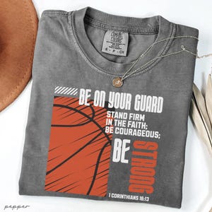 Puede incluir: Una camiseta gris Comfort Colors con un gráfico de baloncesto y el texto "BE ON YOUR GUARD, STAND FIRM IN THE FAITH; BE COURAGEOUS; BE STRONG" en blanco y naranja. Un collar dorado descansa sobre la camiseta.