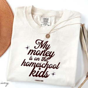 May include: Ivory-colored Comfort Colors t-shirt with the text "My money is on the homeschool kids" in a dark brown script. A gold necklace with a pendant is draped on the shirt.