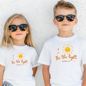 Op de afbeelding: Twee kinderen dragen zonnebrillen en witte t-shirts. De shirts hebben een lachende zon met de tekst "Be the light" en het bijbelvers Mattheüs 5:14 in bruine letters. De kinderen staan voor een witte muur.