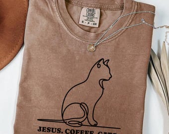 May include: Espresso-colored t-shirt with a black line drawing of a cat and the text "JESUS. COFFEE. CATS." A silver necklace with a small pendant rests on the shirt. The shirt has a Comfort Colors tag.