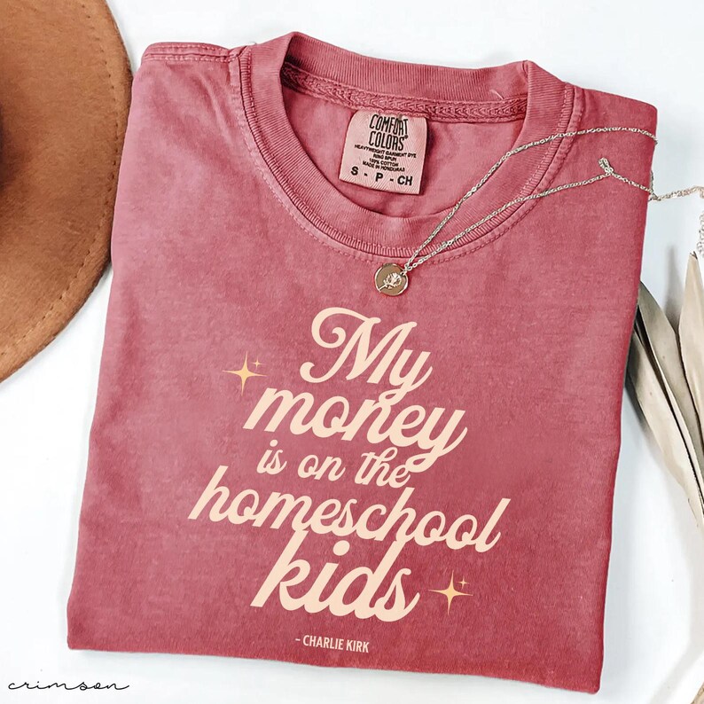 May include: Crimson-colored t-shirt with the text "My money is on the homeschool kids" in a stylized font. The shirt has a crew neck and short sleeves. A silver necklace with a pendant is draped across the shirt.