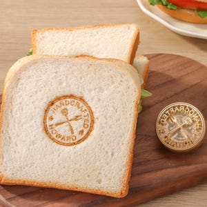 Puede incluir: Una tabla de cortar de madera con un sándwich y una hamburguesa. El sándwich tiene un sello que dice "HANDCRAFTED HOWARD". Un sello redondo con el texto "HANDCRAFTED" también está en la tabla. Una hamburguesa con lechuga y tomate está en un plato blanco.