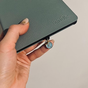 May include: A teal blue notebook with a black zipper and a gold charm with a blue letter K. The notebook has the word "remarkable" embossed on the cover.