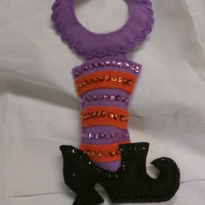 Puede incluir: Colgador de puerta de bota de bruja de fieltro morado con lentejuelas naranjas y moradas. La bota tiene un zapato de fieltro negro con punta.
