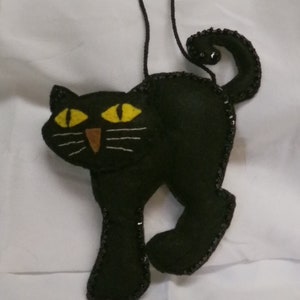 May include: A black cat ornament with yellow eyes and white whiskers. The cat is made of felt and has a beaded outline. A brown string is attached for hanging. The cat's tail is curled.