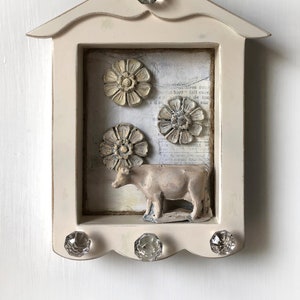 May include: A cream-colored wooden wall-mounted display box with a gabled top. The interior features a printed paper background, three circular floral relief ornaments, and a small cow figurine. Four clear glass knobs are attached to the frame.