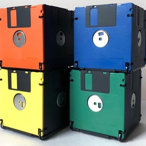 Peut inclure: Quatre cubes de rangement pour disquettes empilés, aux couleurs vives. Les cubes sont orange, bleu, vert et jaune, avec des accents noirs et un moyeu central argenté. Le design est de style rétro et géométrique.
