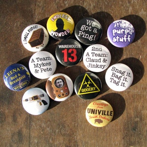 13 one inch buttons show on a wood background. Each has an illustration representing aspects of the series Warehouse 13. One shows Artie on the Farnsworth, one says We got a Ping!, one shows a hazard sign with the word Gooery written inside.