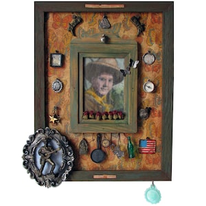 May include: A rectangular shadow box frame with a dark green border and orange butterfly patterned backing. It contains a central portrait of a person in a hat, surrounded by various metal charms, a small skillet, a green bottle, and a US flag.