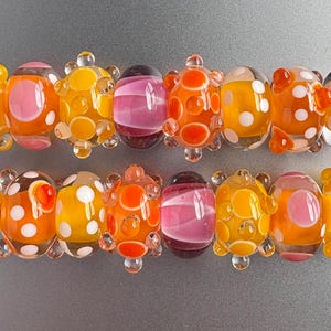 May include: Two rows of glass beads featuring a variety of designs, including translucent orange and yellow spheres with white polka dots, solid pink and purple striped beads, and orange beads with raised circular textures and white accents.