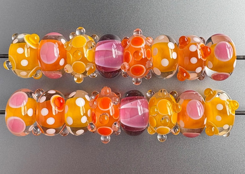 May include: Two rows of glass beads featuring a variety of designs, including translucent orange and yellow spheres with white polka dots, solid pink and purple striped beads, and orange beads with raised circular textures and white accents.