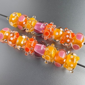 May include: Two strands of glass beads featuring a vibrant color palette of orange, yellow, and pink. The beads are decorated with raised polka dots and circular patterns, with some beads displaying translucent qualities and others showing solid hues.