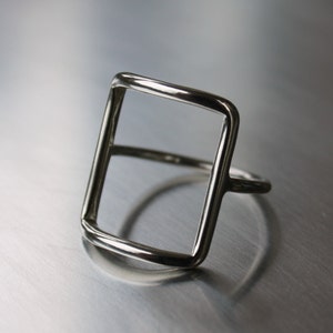 May include: A silver ring with a square shape. The ring is made of thick, smooth metal and has a simple, modern design.