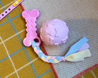 May include: Pink dog toy set featuring a bone-shaped chew toy, a textured ball, and a braided rope with pastel fabric ends. A pink dog collar with yellow and purple accents is also visible. The toys are on a yellow and white plaid blanket.