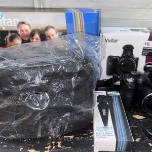 May include: A black Vivitar digital camera with a lens cap, a black camera bag in a plastic wrap, a Vivitar compact tripod in a box, and two Vivitar product boxes. The camera box displays the text 16.1 MP and 12x Power Zoom, while the tripod box shows text.
