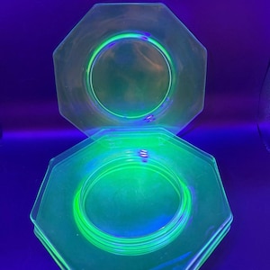 May include: Two octagonal plates made of translucent glass that emit a bright, vivid neon green glow under ultraviolet light. The plates feature a smooth, flat center surrounded by a raised rim, creating a distinct geometric shape and appearance.