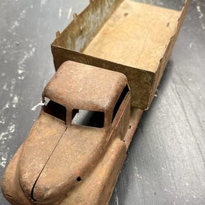 May include: A vintage metal toy dump truck featuring a heavily rusted brown finish across the cab and hood. The truck has an open-top cargo bed with vertical side panels and a rectangular shape, showing significant signs of age and oxidation.