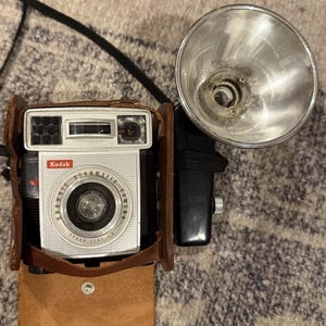 May include: A silver Kodak Brownie Starmatic camera with a black and silver flash attachment, housed in a brown leather case. The camera face features the text Kodak, Brownie Starmatic Camera, and Kodak Lens f/8. The flash unit is circular and metallic.