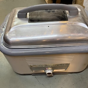May include: A vintage electric roaster oven with a cream-colored base and a metallic silver lid featuring a gray handle. The front includes a horizontal ribbed design, a rectangular metallic control panel, and a circular temperature dial knob.