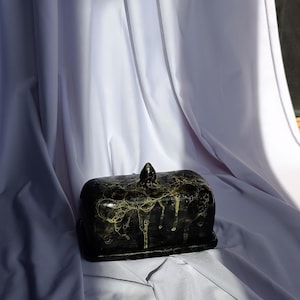 Puede incluir: Una mantequera negra y dorada con tapa y un pequeño pomo en la parte superior. El plato tiene una base rectangular y una parte superior redondeada. Los detalles dorados gotean por los lados. Un artículo de cocina decorativo.