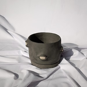 Può includere: Un vaso di ceramica grigio scuro con un'ampia apertura e una piccola base rotonda. Il vaso ha due piccole maniglie sporgenti e una singola apertura di forma ovale. Il vaso è appoggiato su una superficie bianca.