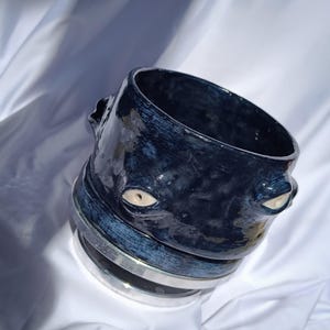 Puede incluir: Una taza de cerámica azul oscuro con un diseño único. La taza presenta dos detalles en forma de ojo y una banda plateada alrededor de la base. El acabado brillante refleja la luz, resaltando la textura y la forma de la cerámica hecha a mano.