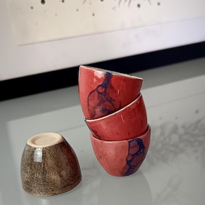 Puede incluir: Un juego de cuatro tazas pequeñas de cerámica. Tres tazas están apiladas verticalmente, con un esmalte rojo brillante y patrones orgánicos azul oscuro. La cuarta taza está separada y presenta un acabado marrón texturizado y moteado.