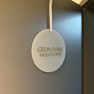 May include: A round, frosted-white sign with the text "GELIN EVINE. HOSGELDINIZ" in gold lettering. The sign is adorned with small, white, pearl-like embellishments and hangs from a white ribbon. The sign is hanging on a gray wall.