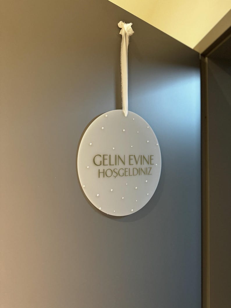 May include: A round, frosted-white sign with the text "GELIN EVINE. HOSGELDINIZ" in gold lettering. The sign is adorned with small, white, pearl-like embellishments and hangs from a white ribbon. The sign is hanging on a gray wall.