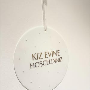 May include: A white, round decorative sign with the text "KIZ EVINE HOŞGELDINIZ" in gold lettering. The sign is suspended by a white string and features small, raised dots around the edge.