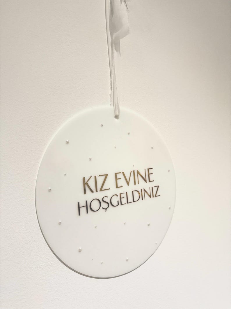 May include: A white, round decorative sign with the text "KIZ EVINE HOŞGELDINIZ" in gold lettering. The sign is suspended by a white string and features small, raised dots around the edge.