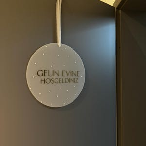 May include: A round, frosted acrylic sign with the text "GELIN EVINE. HOSGELDINIZ" in black lettering. The sign is adorned with small, scattered embellishments and hangs from a white ribbon. The sign is hanging on a dark gray wall.