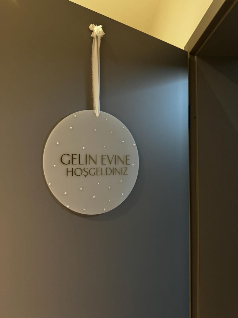 May include: A round, frosted acrylic sign with the text "GELIN EVINE. HOSGELDINIZ" in black lettering. The sign is adorned with small, scattered embellishments and hangs from a white ribbon. The sign is hanging on a dark gray wall.